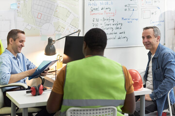 Middle aged Caucasian men and Black man discussing project details in office, foreman wearing...