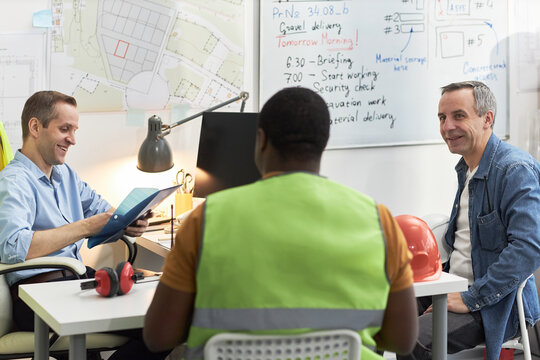 Middle aged Caucasian men and Black man discussing project details in office, foreman wearing safety vest sitting with colleagues near construction plans and safety helmet on table
