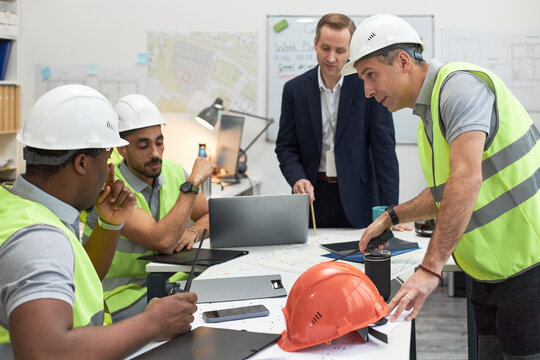 Middle aged Caucasian man in suit supervising group of young adult multiethnic men in safety vests and helmets, discussing construction plans in office setting with blueprints and laptops