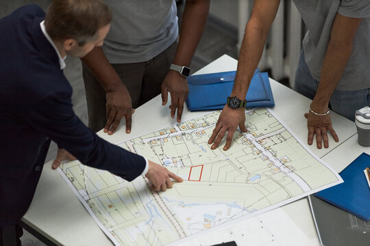 Middle aged Caucasian man and two young adult men, one Black and one possibly South Asian, reviewing architectural blueprint on office table, foreman pointing at building plan