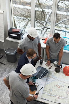 Vertical of group of middle aged men including Caucasian and Black foreman standing around office table discussing construction plans with laptop, blueprints, coffee pot and safety helmets visible