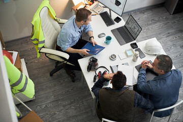 Three middle aged men including Caucasian foreman discussing construction project at office desk,...