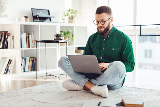 Man Using Laptop Computer Sitting On Floor Wearing Glasses, Working Online Browsing Internet And Typing At Home. Distance Job, Technology And Freelance Career Concept