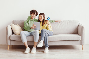 Obraz premium Millennial Asian couple with credit card and digital tablet sitting on sofa, ordering goods on internet, buying things in web store at home, copy space. Online shopping, remote banking concept