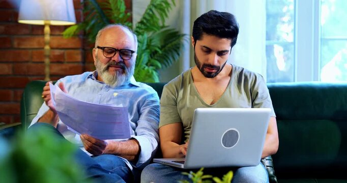 Indian retired father and young son working together on laptop and smartphone at home, reviewing financial report, share trading data and online business documents while discussing profit and loss