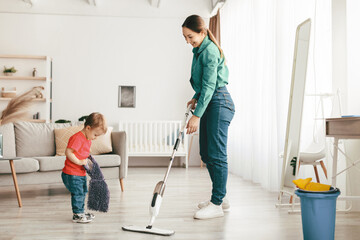 Little helper. Mother and her toddler son doing the cleaning together in living room interior, kid boy giving mom a rag while woman washing the floor with mop