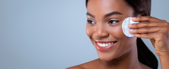 A woman uses a cotton pad to clean her face as part of her morning skin care routine. She smiles while looking in a mirror. The setting is bright with a light blue background. © Prostock-studio