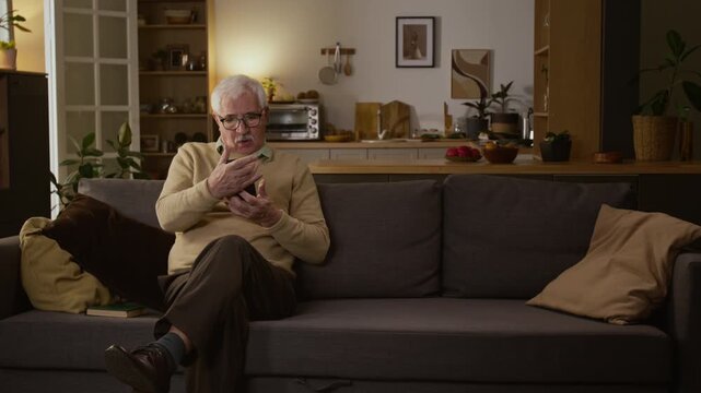 Full shot of elderly 80-year-old Caucasian man relaxing on couch at home, reading book, seeing bank account login attempts on smartphone by cyber scammers, viewing with alarm and confusion