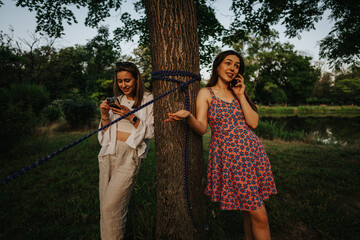 Two women in a park share a moment, one on her phone and the other chatting beside a tree bound...