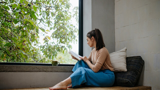 Hygge moment for beautiful woman sitting by a large window with a book. Woman is enjoying time for herself, reading.