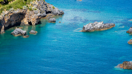 Aerial view of vibrant, crystal-clear turquoise Mediterranean waters. The image captures rugged rock formations and a reef partially submerged along the coastline. © Stefano Tammaro