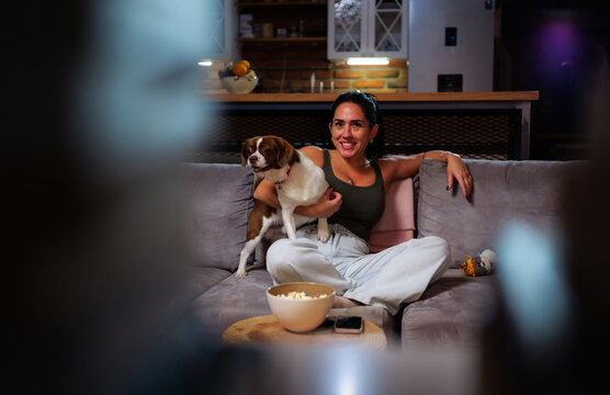 Young woman smiling on a cozy sofa, cuddling her dog and eating popcorn while watching a movie at home, relaxed evening bonding with her pet companion