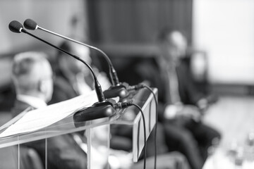 Business conference podium with microphones, executives at round table meeting in background,...