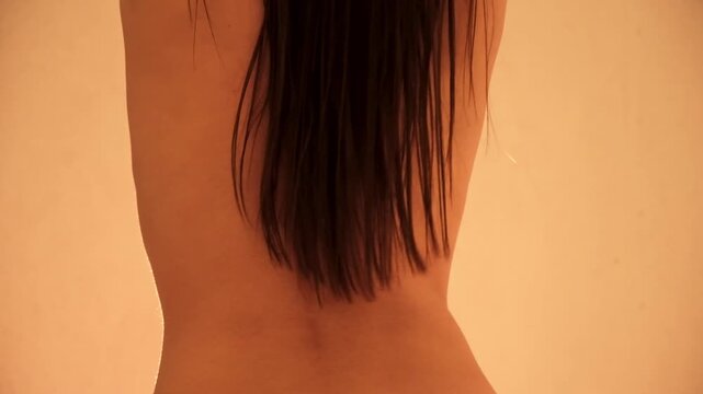 Rear view on back of young naked sexy dancing woman with long dark hair cascading down. Clean and smooth skin. Studio lighting. Evening body bliss. Arousal and lust. Intimate and caring gesture. 