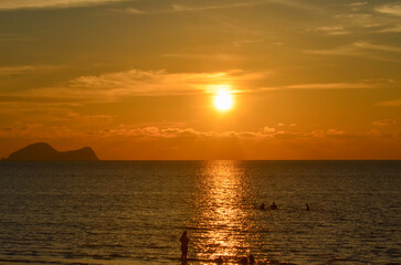 Golden Sunset Over Ocean Horizon with Distant Island.A wide shot of a glowing orange sunset reflecting on the calm sea surface with the silhouette of a tropical island in the background.