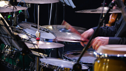 Close-up of a drummer's hands playing a drum set. Motion blur on the drumstick hitting a cymbal captures the energy of a live performance. Stage lighting highlights instruments on a dark background © VeNN