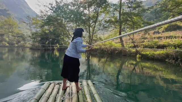 A woman poles a bamboo raft on on a river in Hang Than Lim, Yen Minh, Ha Giang, Vietnam. She is using a long pole to push the raft forward, enjoying the scenery and the peacefulness of the river.