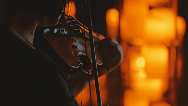 Close-up of a violinist performing in a dark, intimate setting. The musician is silhouetted, lit by a warm, golden glow from blurred candlelight, creating a moody and atmospheric bokeh effect