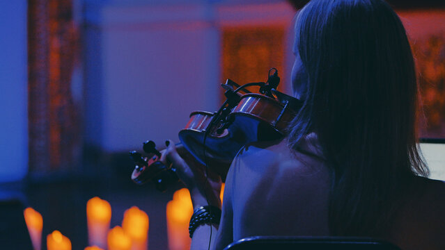 Over-the-shoulder shot of a woman playing the violin at an intimate concert. Dramatic blue and warm orange light from candles creates a moody, atmospheric and cinematic feel