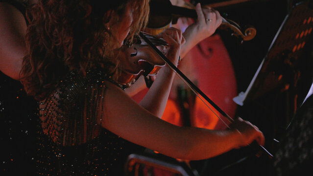 Close-up of a female musician playing the violin during a classical performance. Warm, dramatic stage lighting highlights her elegant, sparkly dress. Dark, atmospheric mood of a symphony concert