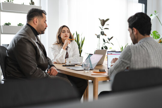 A diverse team of three professionals collaborates around a table, discussing ideas and working on laptops in a bright, modern office setting.