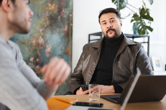 Two men are in a meeting, one is looking at the camera while the other is out of focus. They are at a table with a laptop and notebooks.