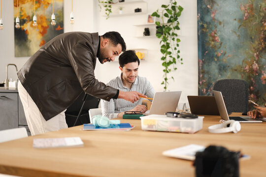 Two men collaborate on a project at a wooden table, one pointing at a laptop screen with a pencil, surrounded by office supplies and modern decor.