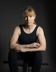 Serious portrait of middle aged woman in fitness attire in studio sitting on gym equipment