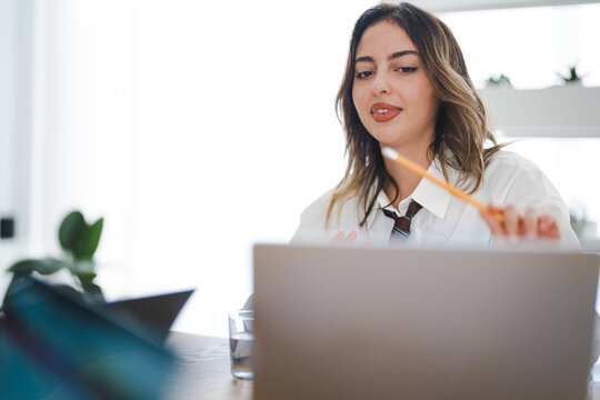 A young woman in a white shirt and tie is working on her laptop, holding a pencil and looking at the screen.