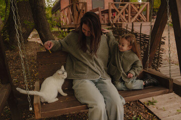 Mom and daughter sitting on a swing with a white cat and playing. A cheerful family on vacation. A wooden bathhouse in the background. People love animals. Relationships between people and animals. 