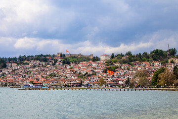 Obraz premium Historic Ohrid town with traditional red-roofed houses climbing a hillside, crowned by an ancient fortress, overlooking a vast lake under a cloudy atmosphere.