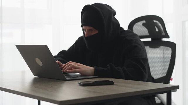 A scammer in a black balaclava with a laptop.
Hacking a laptop password.
Financial fraud.
Hacker in a black mask.