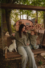 Mom and daughter are sitting with a cat on a swing at a recreation center near the river. A white cat is playing with a girl. Pets are allowed at the hotel. Vertical photo.