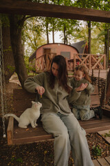 Mom and daughter are sitting with a cat on a swing at a recreation center near the river. A white cat is playing with a girl. Pets are allowed at the hotel. Vertical photo.