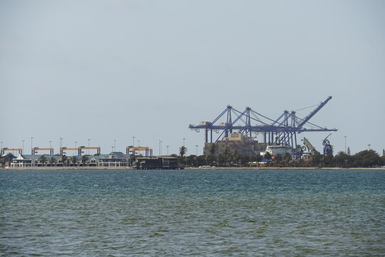 Industrial port of Swakopmund, Namibia, featuring large blue container cranes, stacked shipping containers, and a cargo vessel at the Atlantic Ocean waterfront.