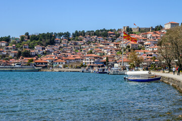 Fototapeta premium A bustling Ohrid waterfront features colorful houses climbing a lush hillside, boats docked, and a Macedonian flag under a clear blue sky.