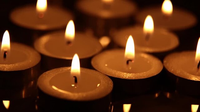 Close-up of tea candles burning indoors at night against a dark background. The flame flickers and the wax cups remain stationary, creating a warm, memorable glow with a bokeh effect.