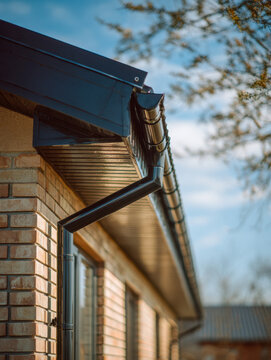 Modern black rain gutter system installed on beige brick house exterior with clear blue sky and soft sunlight on a calm day in suburban area