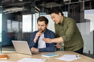 Two diverse business partners discussing work, looking at a laptop screen, and pointing at data...