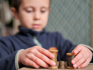 child's hands arranging vintage wooden thread spools. A focused boy in a dark blue fleece jacket interacts with antique sewing supplies, including a metal thimble and wooden bobbins © light