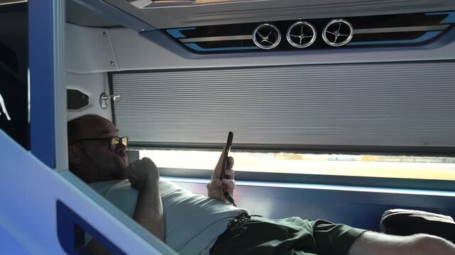 Sleeper Bus Person Reclining On Upper Sleeping Area Focusing On Phone. Male Individual Resting On Upper Sleeping Space Engaging With His Mobile Device While Soft Daylight Lights His Face