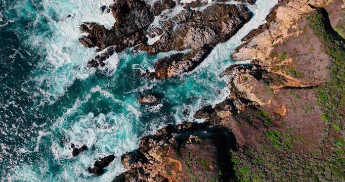 Aerial view of turquoise ocean coves and rocky cliffs. Vertical shot of clear water rushing between coastal rocks and vegetation