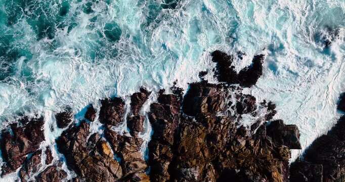 Ocean waves breaking on dark coastal rocks. High angle shot of white surf hitting rocky shore with copy space