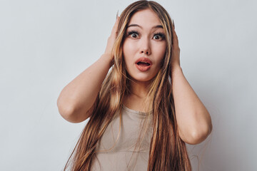 Young woman with long braided hair looks surprised and shocked with hands on head against plain...