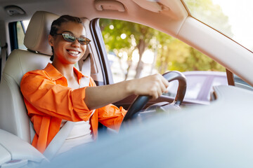 Naklejka premium Young black woman driving car wearing sunglasses, smiling with relaxed expression and enjoying sunny day while commuting safely through the city