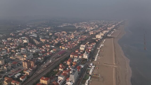 Early morning drone flight at sunrise over Cingoli, Marche, Italy. Light mist drifts above the town while the Adriatic Sea appears on the horizon in soft golden dawn light.