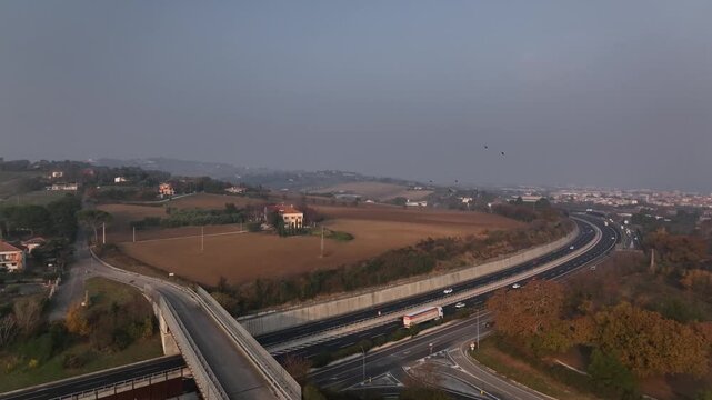 Early morning aerial drone flight over highways near Cingoli, Marche, Italy. Cars speed along the road with the town and Adriatic Sea visible in soft sunrise light.