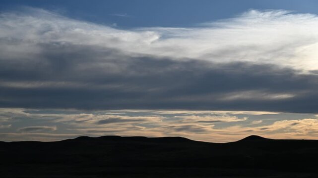 Late-day clouds drift and swirl in a calm time-lapse, gliding above distant rolling hills silhouetted against the fading evening light.