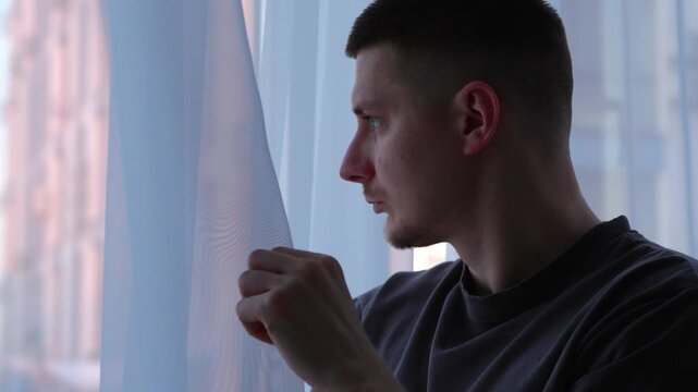 A man near a window.
The man touches the white, transparent tulle hanging on the window.