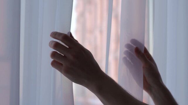 A man stands by the window, touching the sheer curtains.
Interior of a room.
White silk curtains in a cozy room.